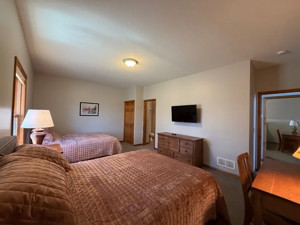 Cottage bedroom with two queen beds, tan quilted bedding, wall-mounted TV, and en-suite entry