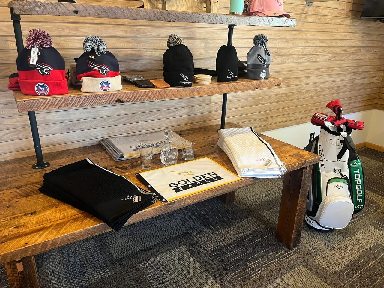 Bench display of Golden Eagle branded beanies, towels, glasses, and a pin flag in the pro shop