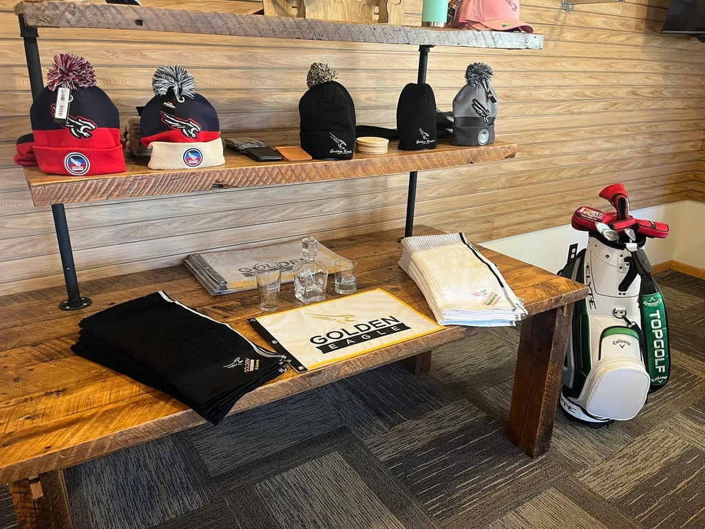 Bench display of Golden Eagle branded beanies, towels, glasses, and a pin flag in the pro shop