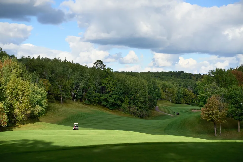 Wide fairway corridor with a single cart in the distance, dramatic clouds drifting over the treeline