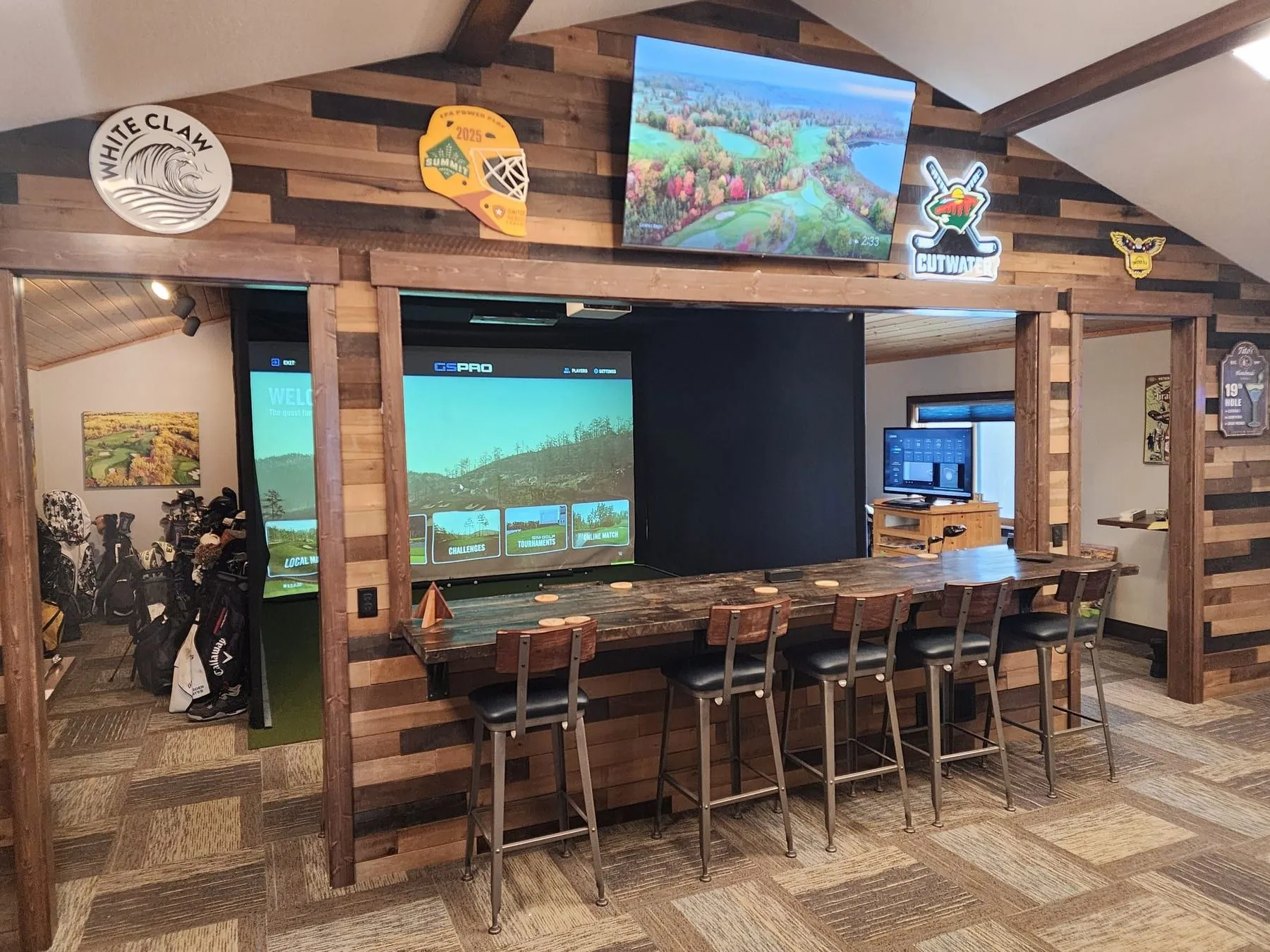 The Golden Eagle simulator lounge with a long bar-height counter and stools facing the GSPro screen, framed by reclaimed wood walls and Northwoods decor