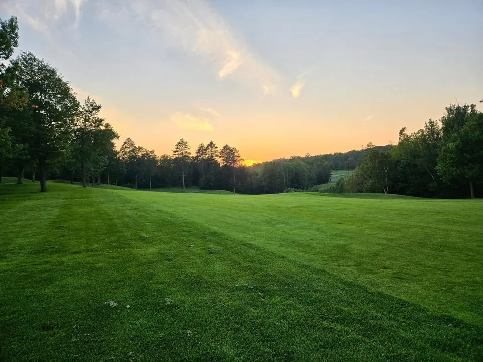 Wide fairway from the tee at sunset, treeline silhouetted against an orange sky