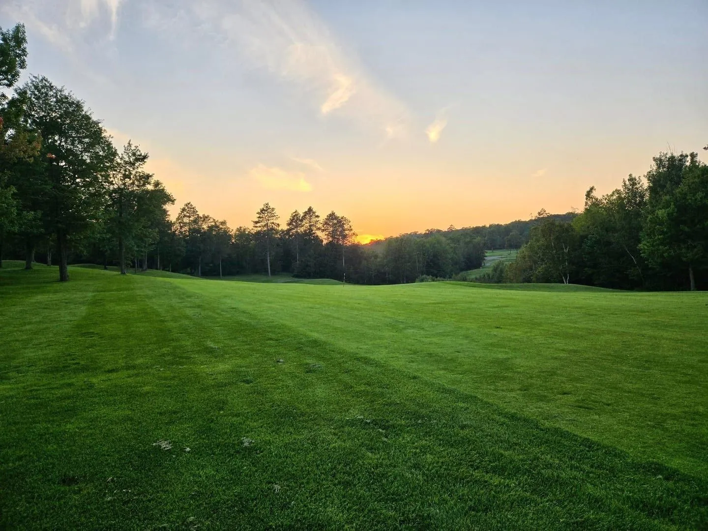 Wide fairway from the tee at sunset, treeline silhouetted against an orange sky