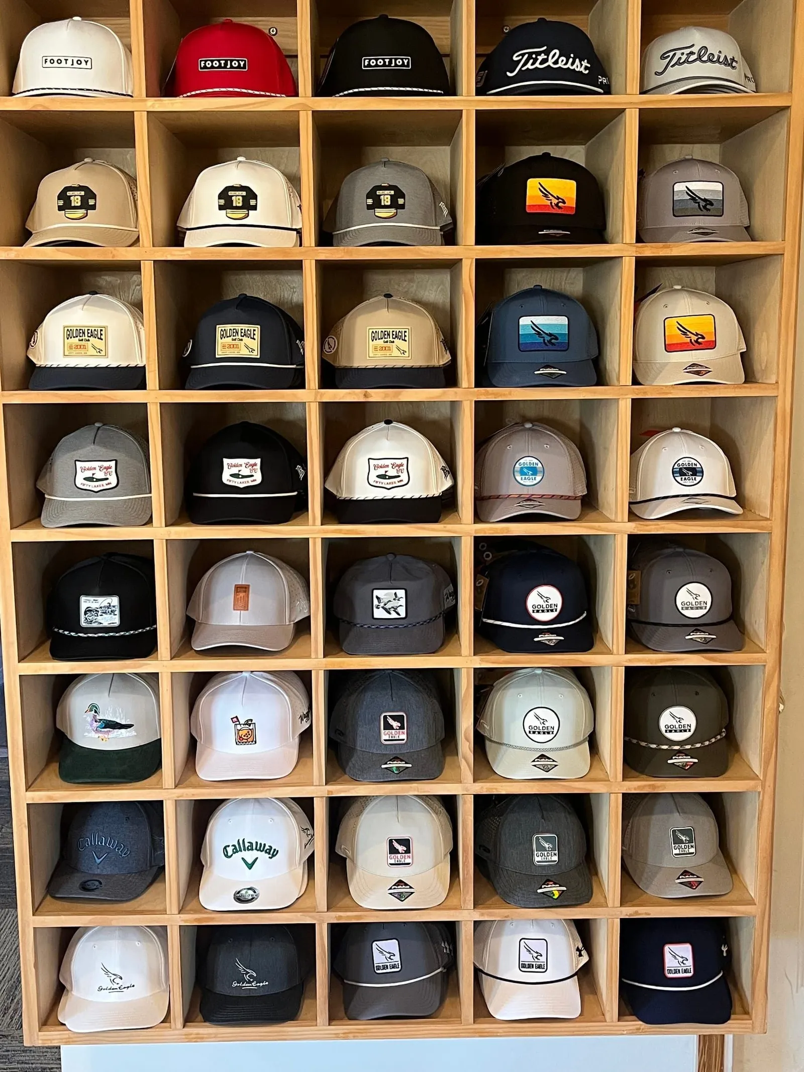 Wall of hat cubbies in the Golden Eagle pro shop stocked with TravisMathew, Branded Bills, FootJoy, Waggle, and Pukka headwear