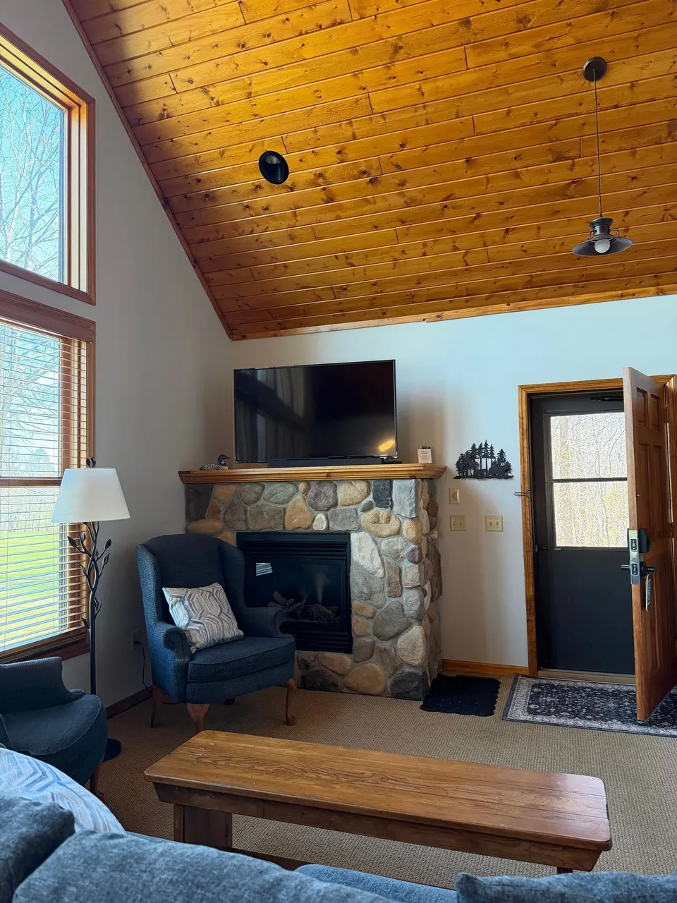 Living room inside the cottage with floor-to-ceiling stone fireplace, leather chair, and vaulted pine ceiling